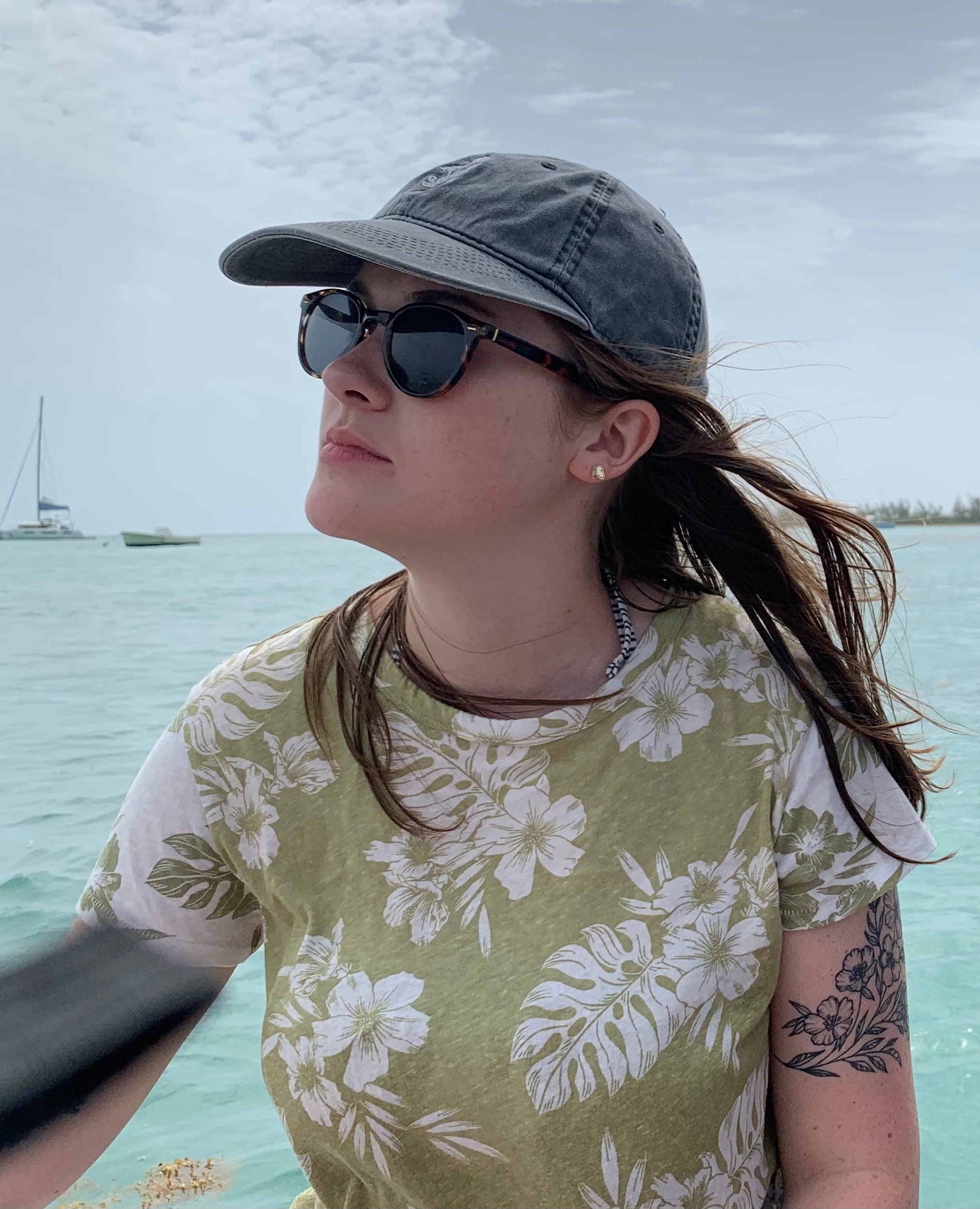 Elliana wears a cap and sunglasses and looks wistfully away while standing on a boat in the backdrop of the ocean.