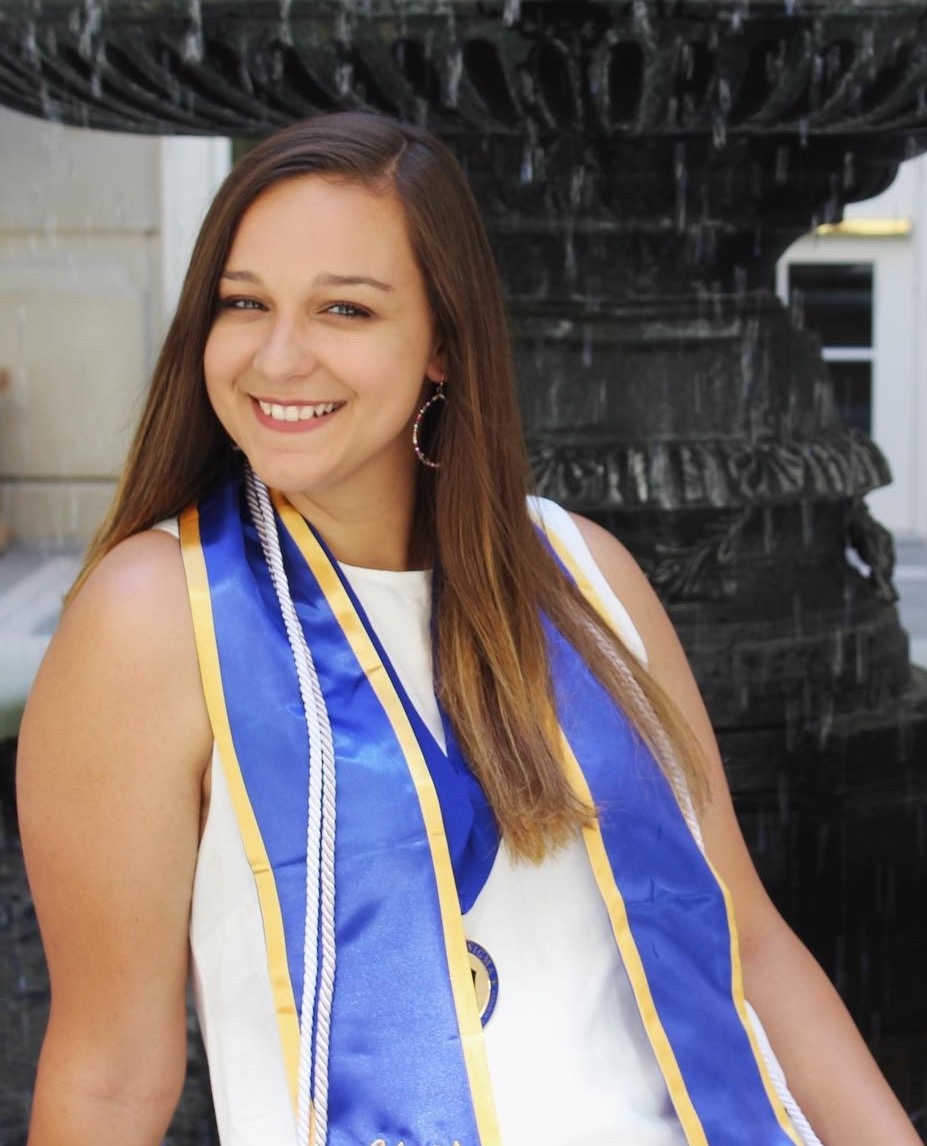Megan smiles in front of a fountain while wearing a white dress and graduation attire.