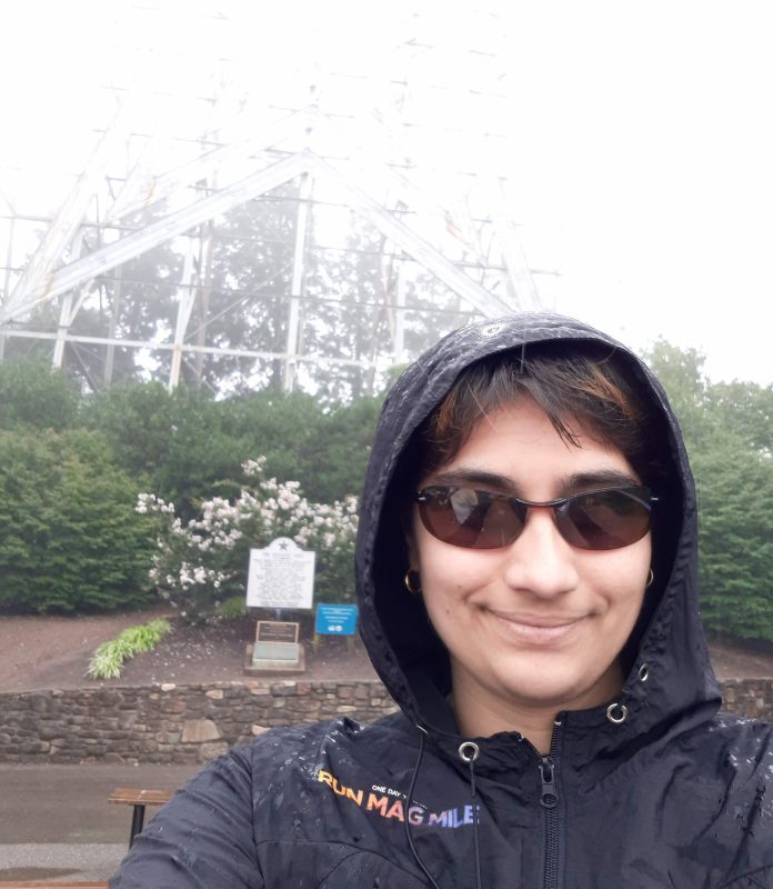 Gates wearing a black rain jacket with the hood pulled up. The jacket says Run Mag Mile in rainbow text on the left shoulder.  She is wearing brown sunglasses, and her short brown hair (her bangs) are poking out under her hood.  Behind her, the base of the Roanoke Star is visible, though obscured in fog.  Because of that, the background is mostly green bushes.